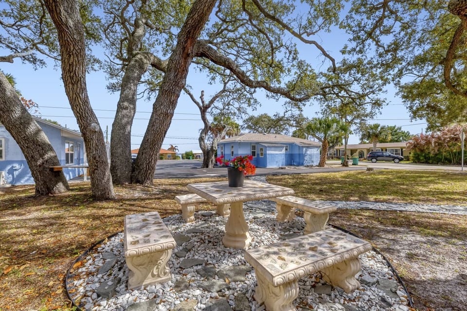 Outdoor seating area with a stone table and benches set beneath leafy trees beside a blue house.