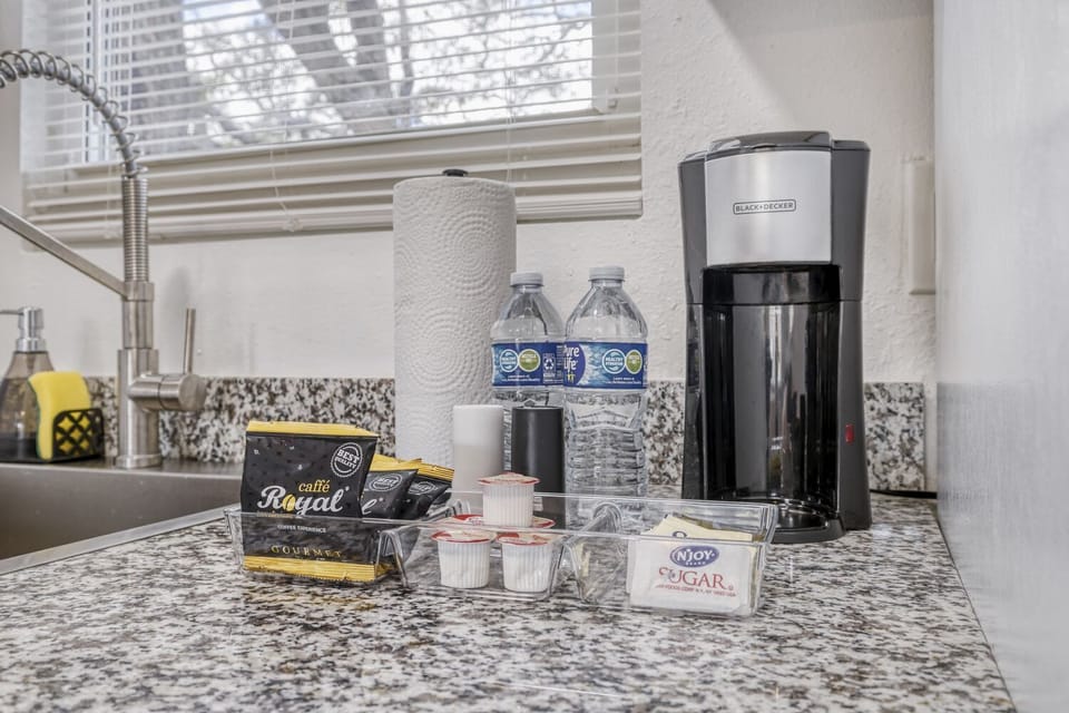 Coffee maker and beverage setup on a granite counter with bottled water, paper towels, and assorted coffee pods.