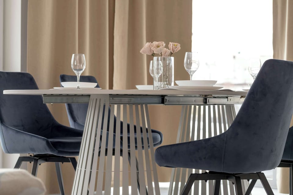 A detailed shot of the dining table shows a minimalist setting with elegant glassware and a small vase of flowers. The stylish blue chairs provide a sharp, attractive contrast against the light interior.