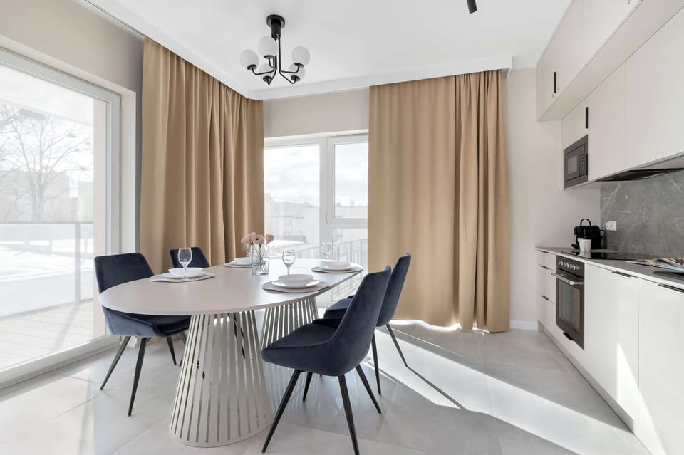 A bright perspective of the dining room showcases the sculptural base of the dining table and its proximity to the kitchen. The flood of natural light from the windows makes this an ideal spot for morning coffee.