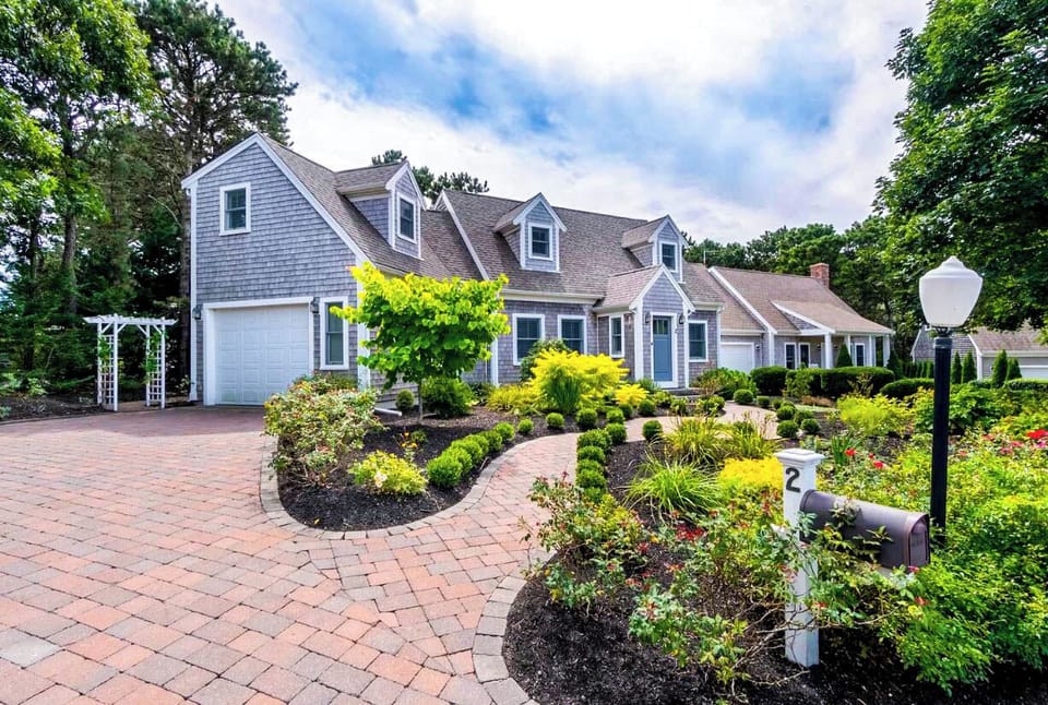Charming Cape Cod-style home with shingle siding, dormer windows, and a spacious layout. Beautifully landscaped gardens and a curved brick driveway create a welcoming, picturesque entrance in a serene, private setting.