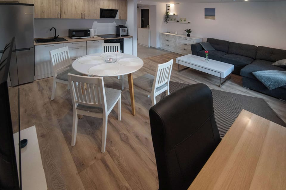 A view of the living area from the hallway's perspective, showcasing the open-plan layout of the apartment. The white round table serves as a central focal point between the kitchen and the living room, highlighting the functionality of the space.