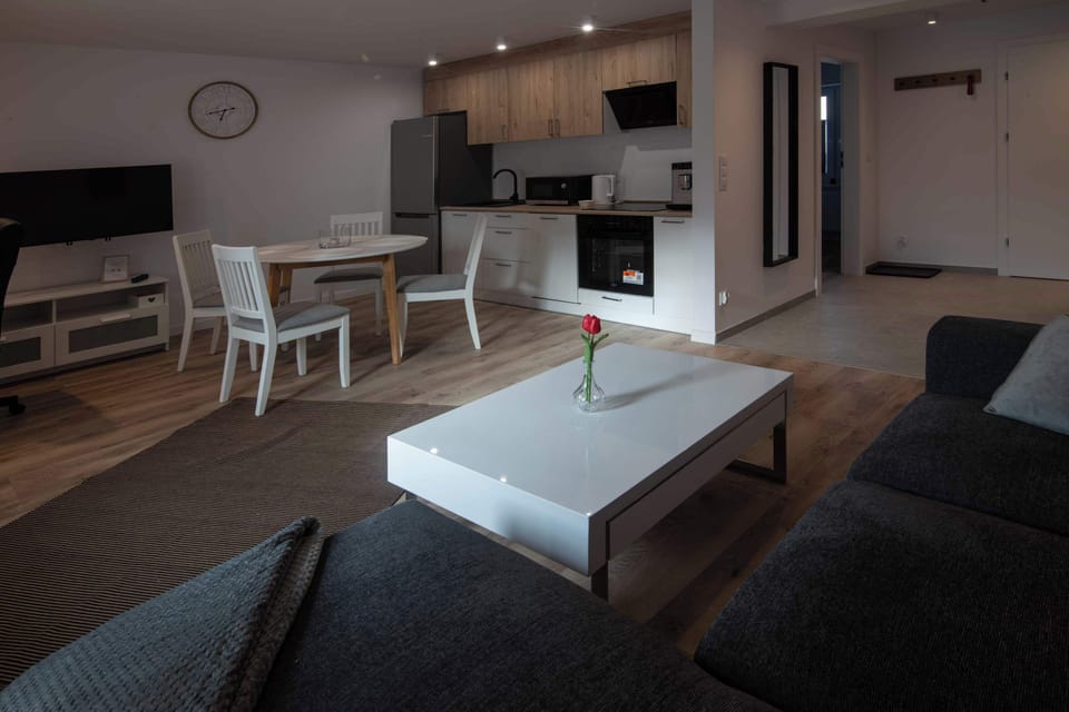 A spacious living room connected to the kitchen, furnished with a dark sofa and a white coffee table. The interior is well-lit, and the wood-like flooring seamlessly connects the lounge area with the dining section.