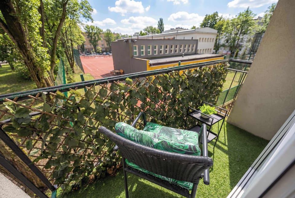 A small balcony surrounded by lush greenery and trees. A narrow outdoor table with chairs is arranged along the railing. The view overlooks neighboring buildings partially hidden by foliage.