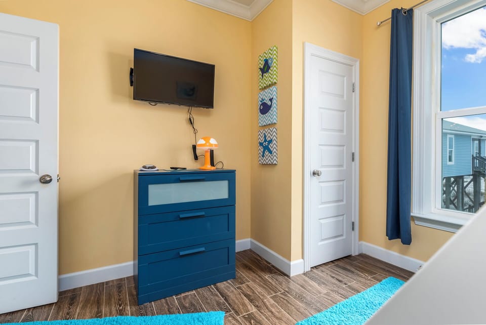 Your bright bedroom features a mounted flat-screen TV and stylish blue dresser, creating the perfect space to unwind after beach adventures.