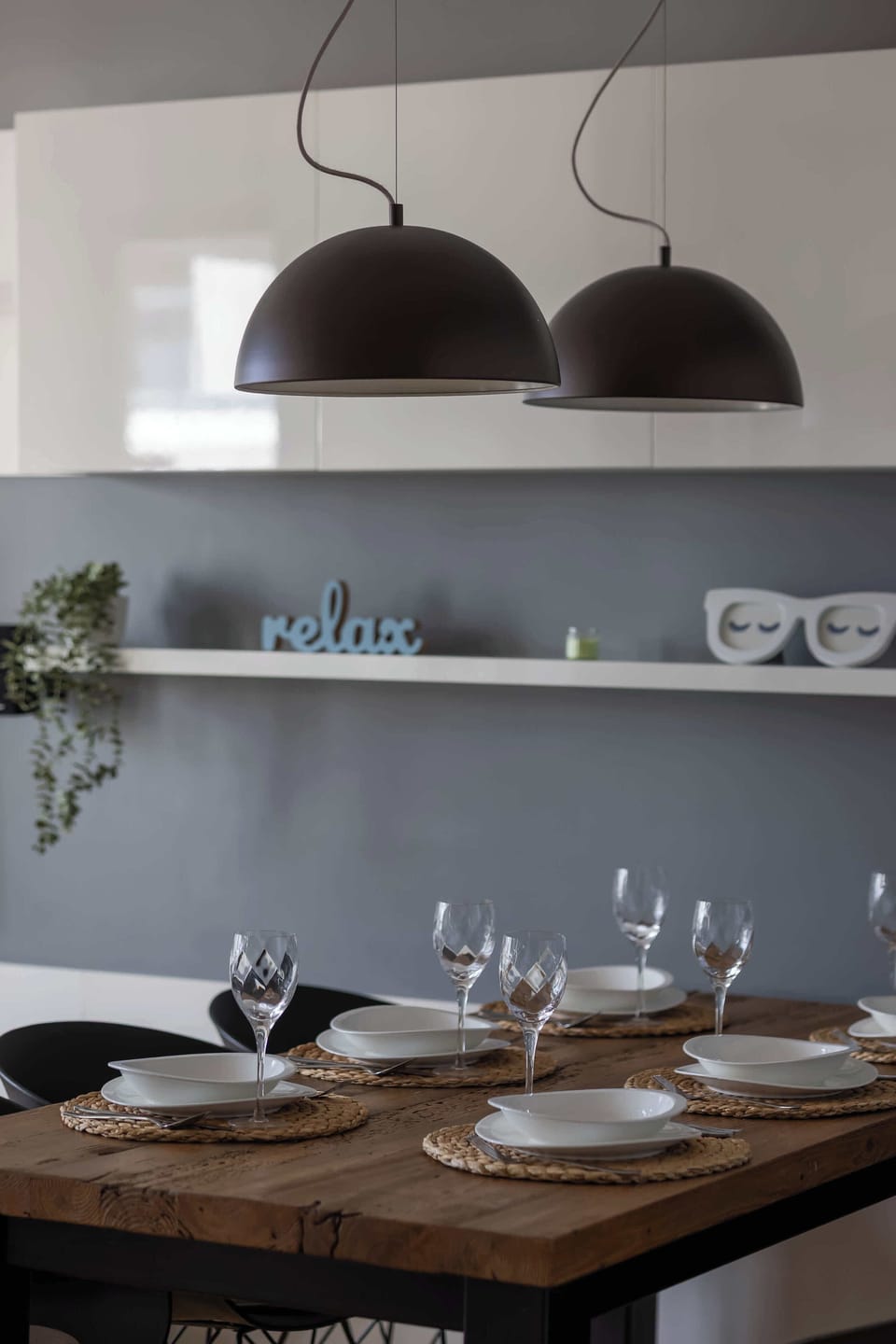 A dining table detail shows a neatly set wooden table with wine glasses and plates. Two large black dome-shaped pendant lights hang low over the surface for focused lighting.