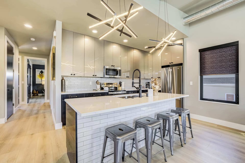 The kitchen features a large island with a classic subway tile aesthetic and industrial-style stool seating. Geometric linear lighting illuminates the professional-grade stainless appliances and high-gloss cabinetry.