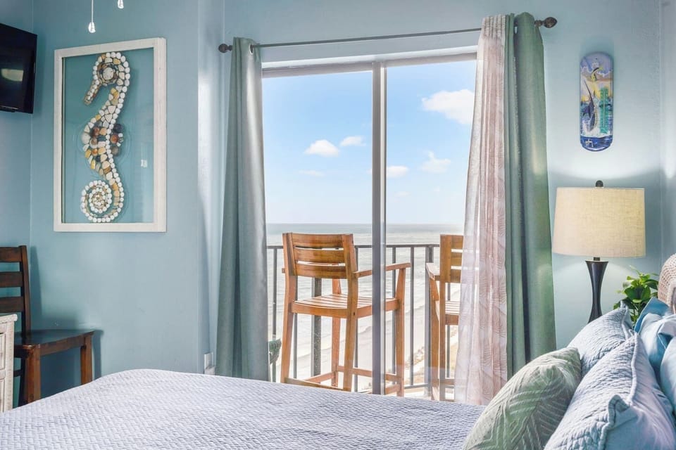 Cozy bedroom with plush queen bed, Smart TV, dresser and closet for clothing storage, beach-inspired décor, and sliding glass door access to the balcony with beautiful ocean views.