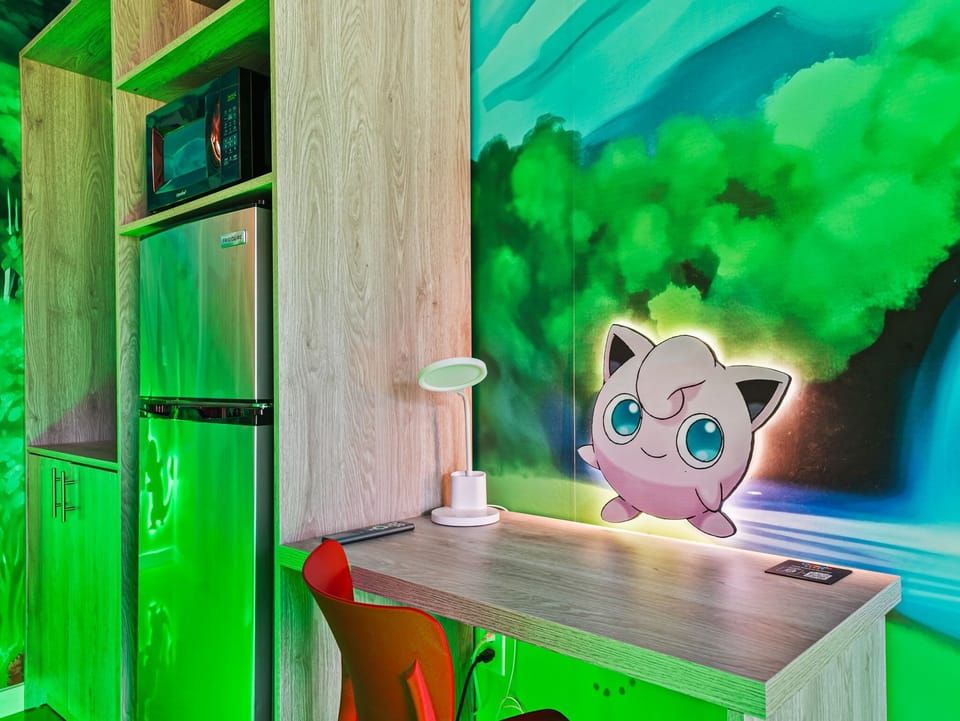 Compact kitchenette with microwave and fridge next to a playful jigglypuff mural and workspace.