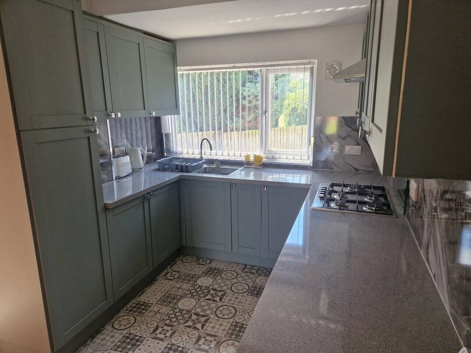 Fitted kitchen with sage-green units, a gas hob, stone-effect worktop, patterned floor tiles, and a window to the side.