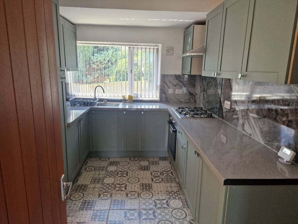 Sage-green kitchen showing dark worktops, a window with a view to the rear garden, and patterned tile flooring throughout.