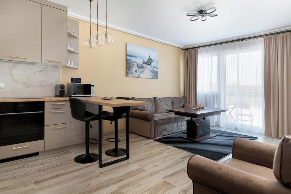 The bright living room opens up to a balcony through large glass doors, offering an airy feel. The combination of beige walls and wooden furniture creates a warm, inviting ambiance.
