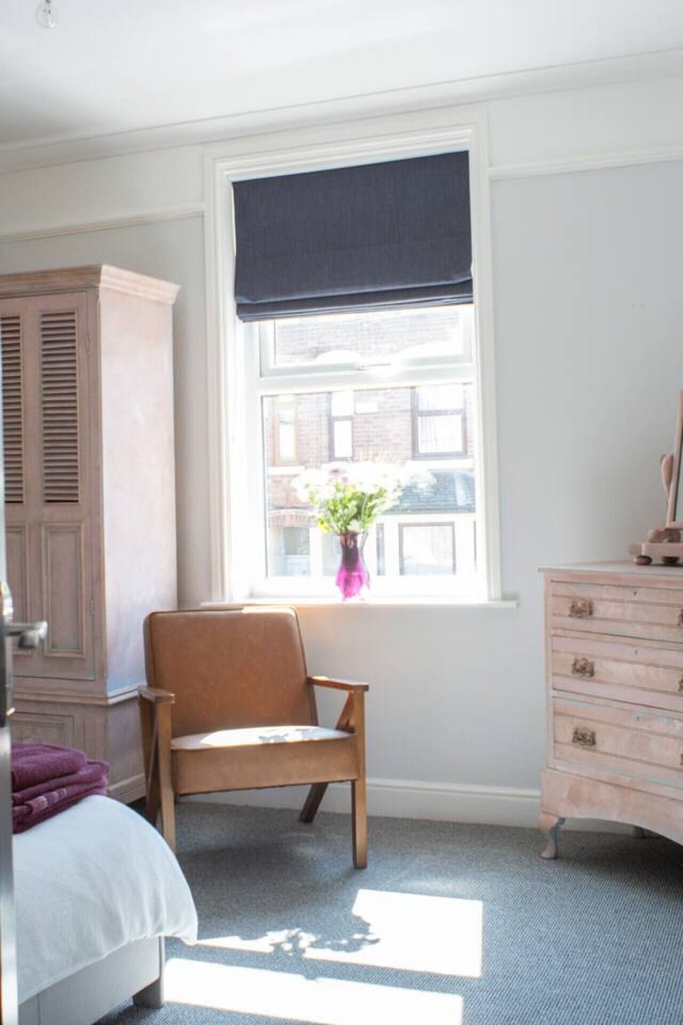Main bedroom in bright daylight with the louvred wardrobe, leather armchair, pink chest of drawers, and a vase of flowers on the windowsill.