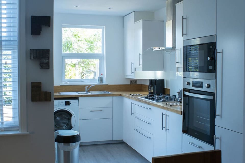 White gloss fitted kitchen with built-in single oven, gas hob, extractor hood, microwave, washing machine, and a window, showing the full run of floor and wall units.