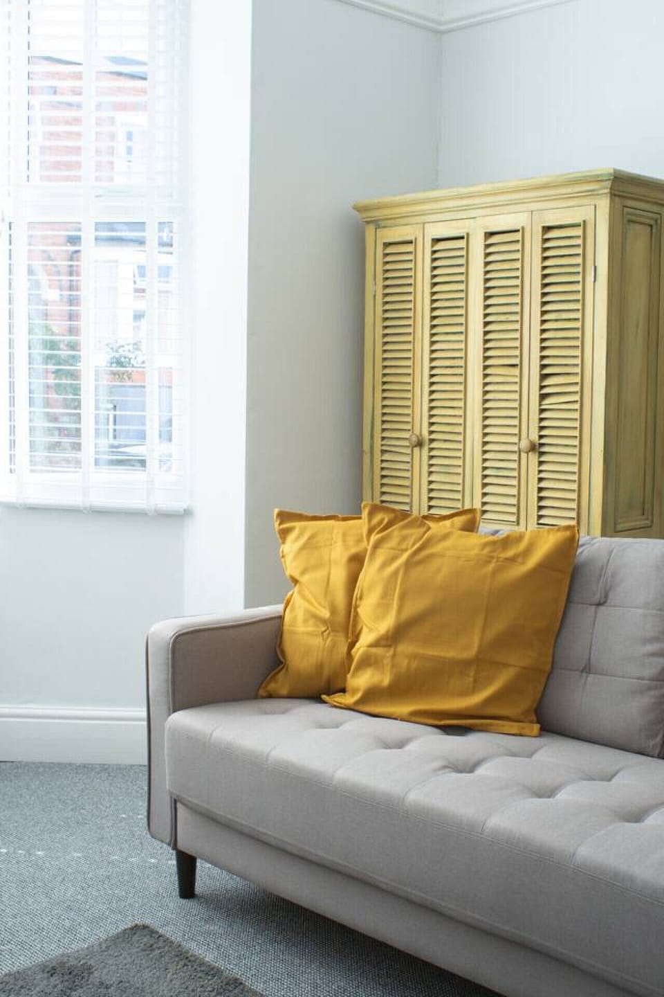 Grey sofa with yellow cushions alongside a yellow painted louvred-door cabinet and a plantation-shutter window in the sitting room.