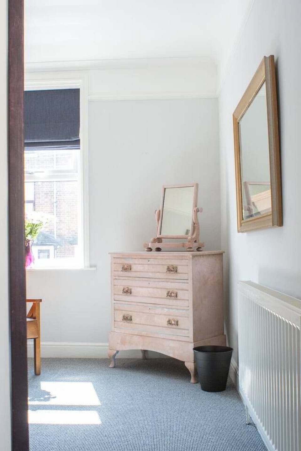 Bedroom with a blush-painted chest of drawers, a large gold-framed mirror above, a standing dressing mirror, and a window with venetian blinds.