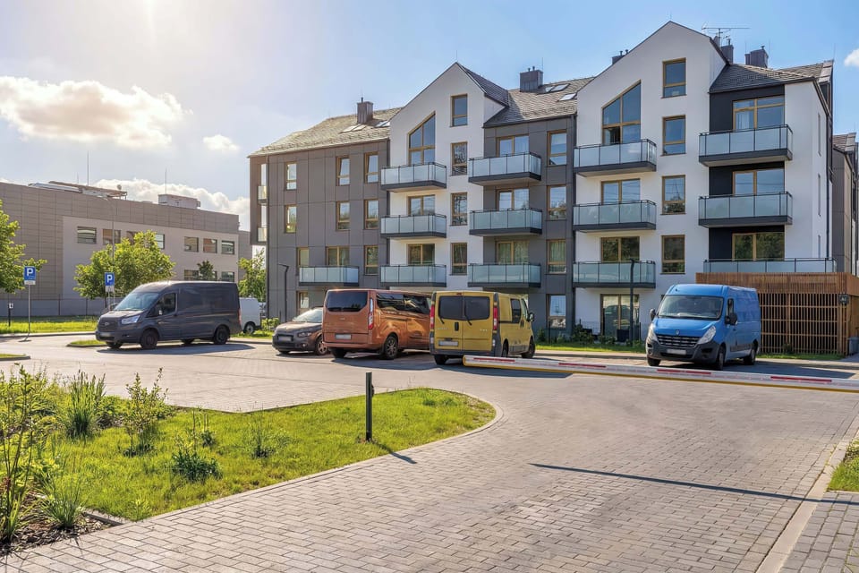 The modern apartment building stands out with its clean facade, balconies, and large windows. The surrounding area includes paved walkways, parking spaces, and neatly maintained greenery.