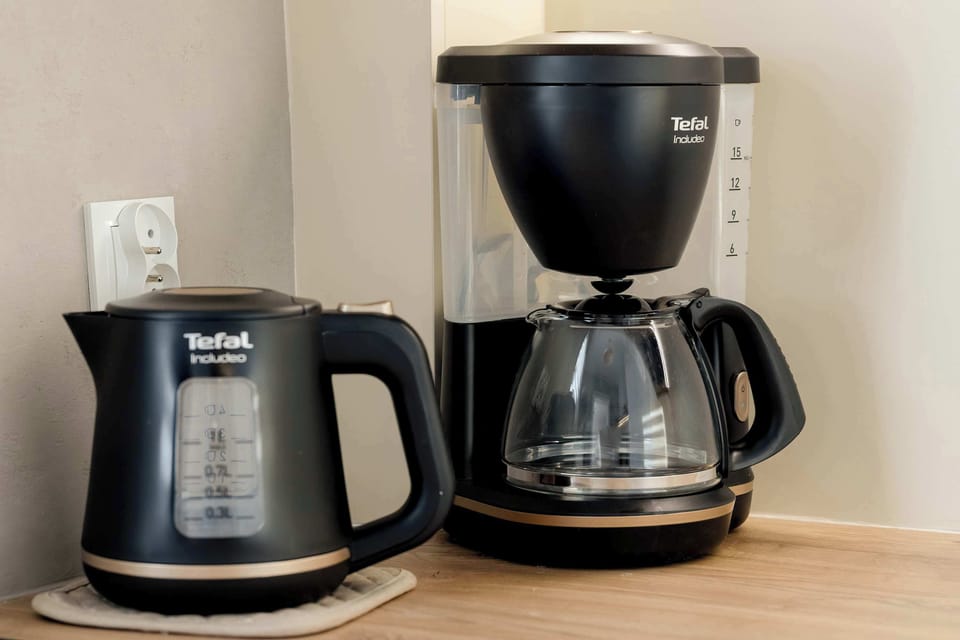 A coffee station set on a kitchen counter featuring a drip coffee maker and an electric kettle. The appliances are black and neatly arranged. The neutral backsplash complements the setup.