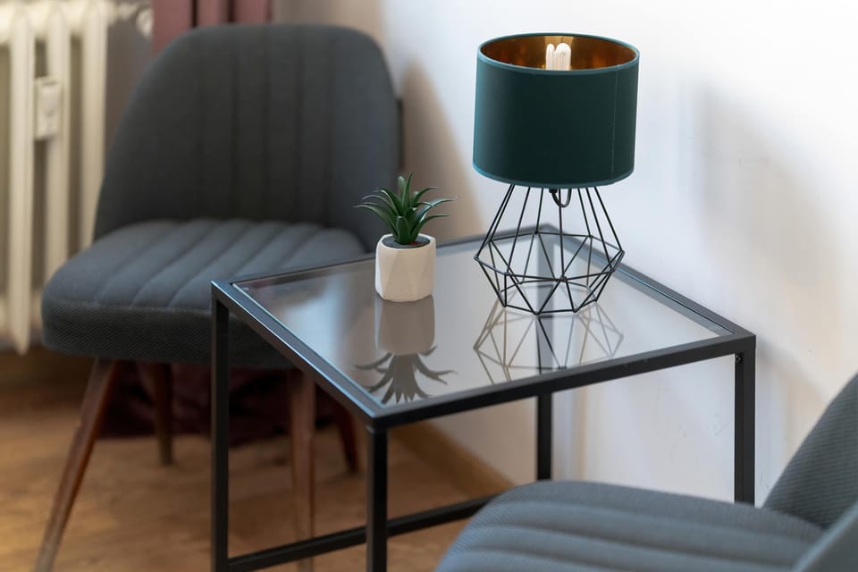 A small seating corner with a gray upholstered chair placed next to a table with a lamp and decorative plant. The corner feels like a quiet reading or relaxation space. Soft lighting and neutral tones make it inviting.