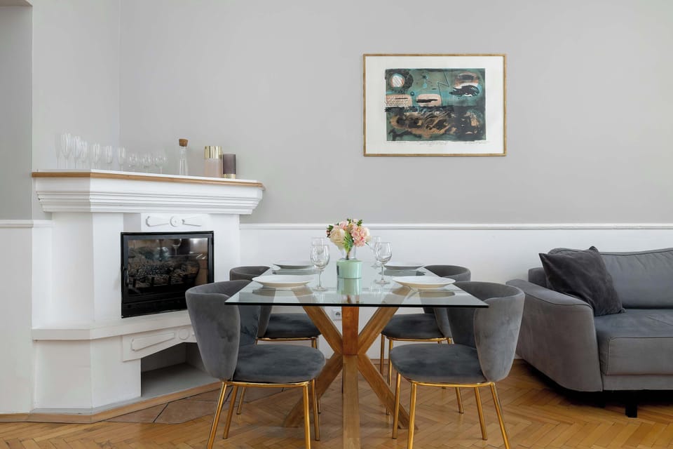 A dining table set with glassware positioned in front of a classic white fireplace mantle. The contrast between the modern furniture and traditional architectural details is striking.