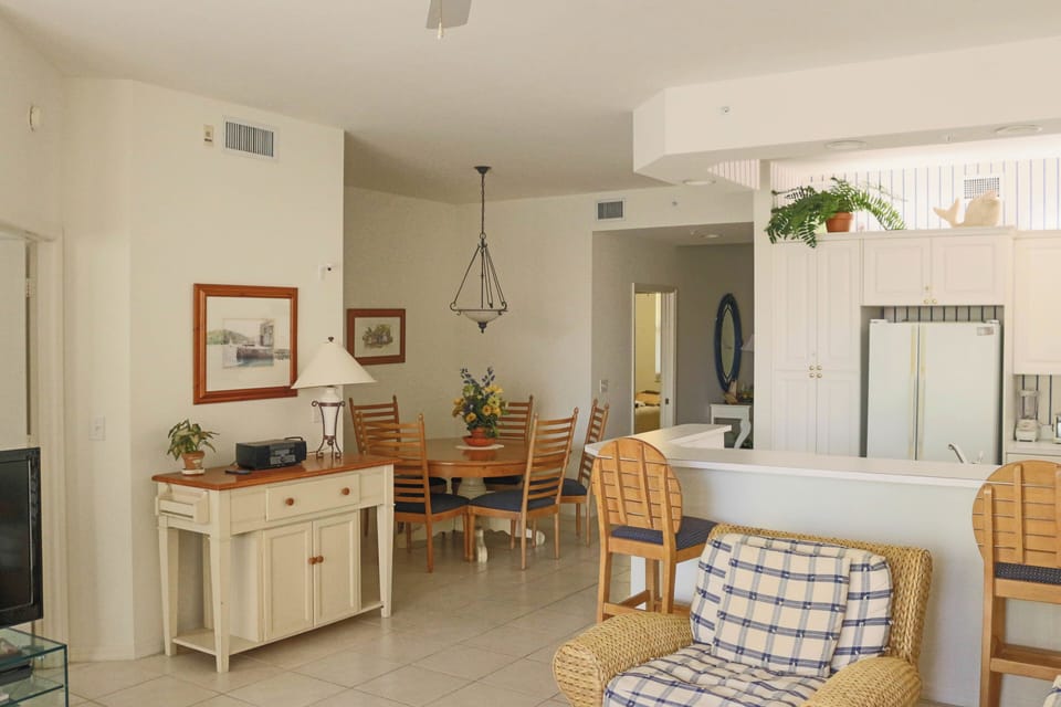 Relax in your bright, open-concept living space featuring coastal charm with wicker seating, warm wood furnishings, and seamless kitchen access.