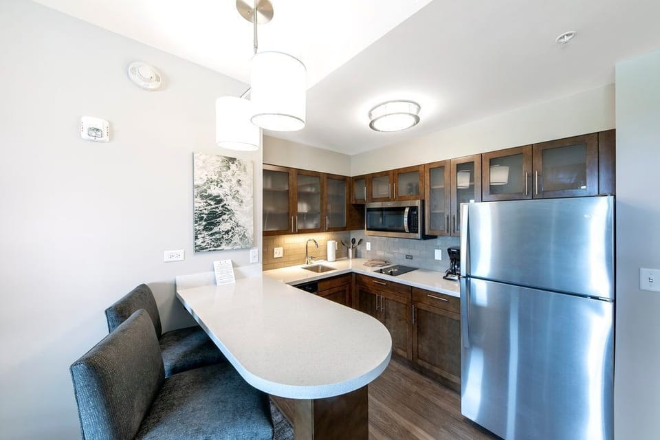 The kitchen features wooden cabinets, a stainless steel refrigerator, a microwave, and a breakfast bar with two cushioned chairs.
