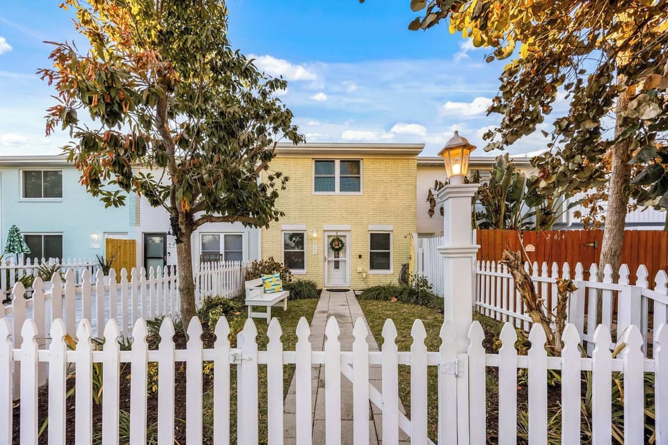 Charming yellow brick townhouse with a white picket fence and inviting front yard seating.