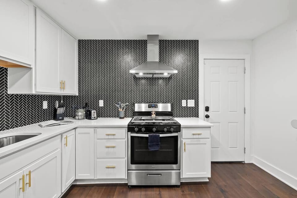 Sleek, fully equipped kitchen featuring white cabinetry, quartz counters, and gold accents. Highlights include a stainless steel stove/oven and a striking black herringbone backsplash. Perfect for preparing any meal.