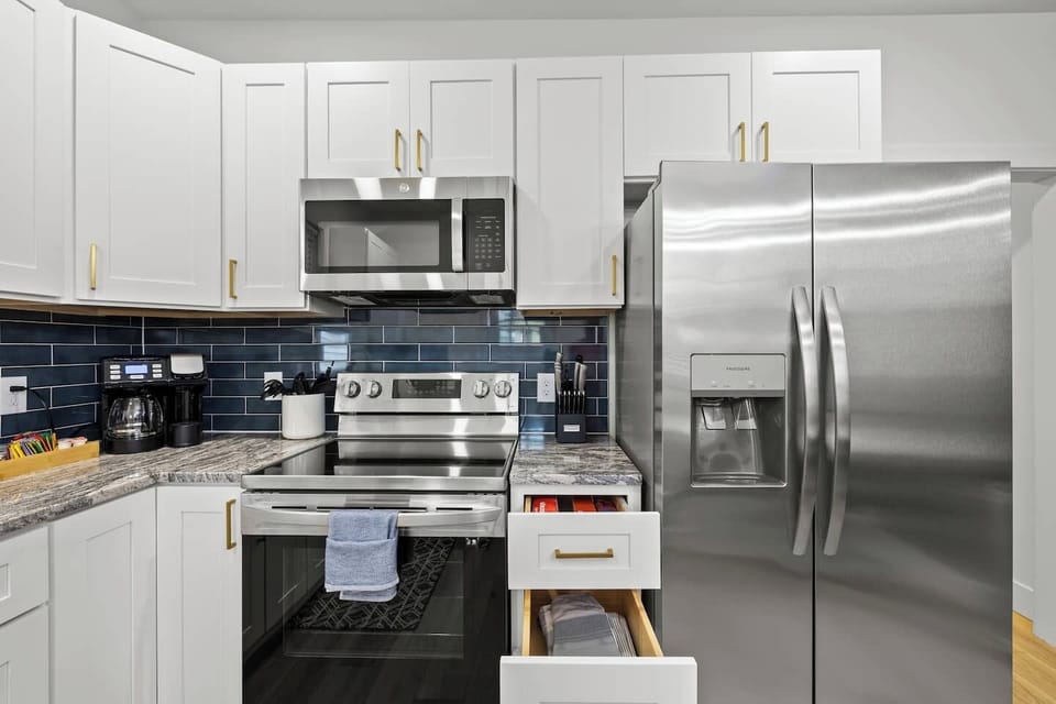 Fully updated kitchen featuring white cabinetry with gold hardware, granite-style countertops, and a striking blue subway tile backsplash. Equipped with stainless steel appliances, including a side-by-side refrigerator, electric range, and microwave.