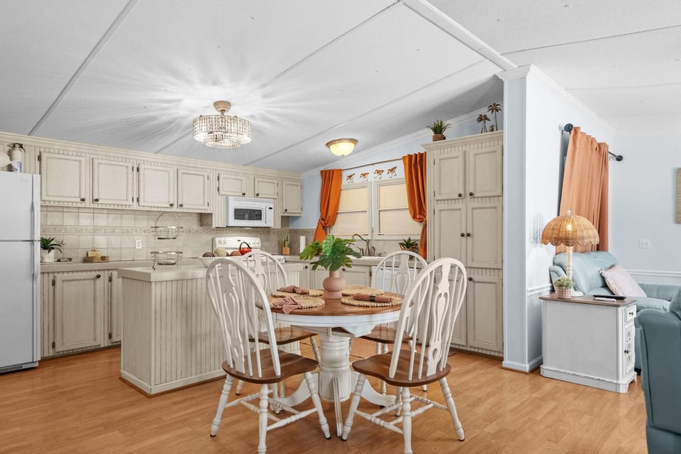 Dining area with round wood table and white spindle-back chairs, adjacent to a cream-toned kitchen with tile backsplash.