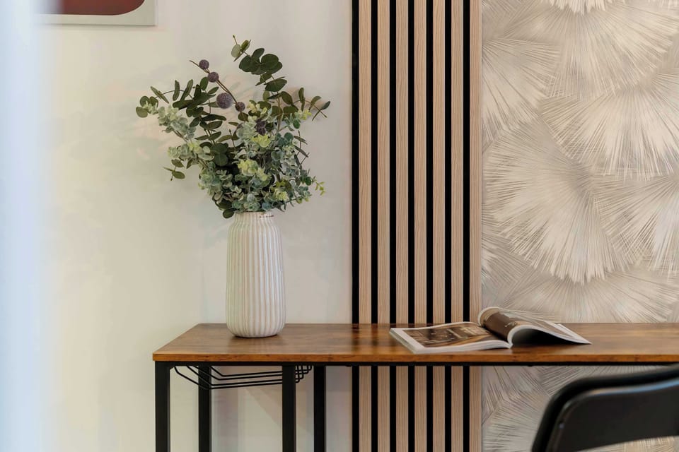 A decorative corner with a slim console table, a vase with green branches, and a textured wall panel behind it.
