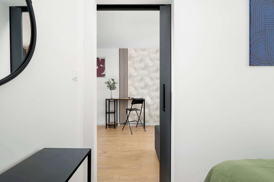 A hallway view with a round mirror on the wall, leading toward a brighter room with light wood flooring.