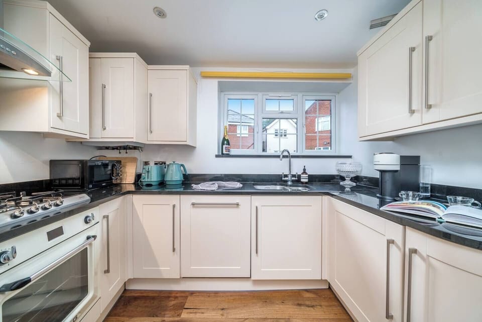 Bright and fully equipped U-shaped kitchen with cream cabinetry, black countertops, and wood flooring. A large window over the sink provides natural light, while modern appliances and teal accents add charm and functionality to the space.