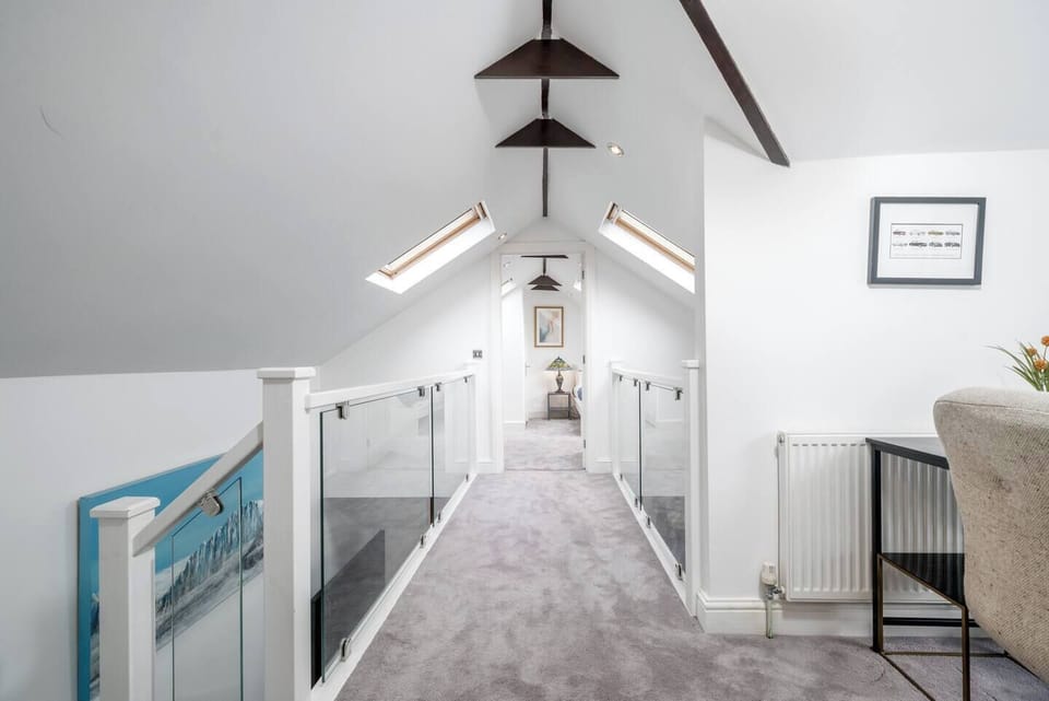 Bright loft hallway with skylights, vaulted ceilings, and exposed beams. Glass balustrades line the walkway, creating an open and airy feel. Soft carpeting adds comfort while leading to a cozy room at the end of the corridor.