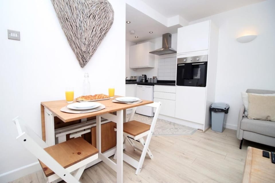 A modern kitchen and dining nook with sleek white cabinetry, built-in oven, and cozy wooden table set for two. A woven heart wall décor adds charm, while the adjoining living area creates a warm, inviting atmosphere.