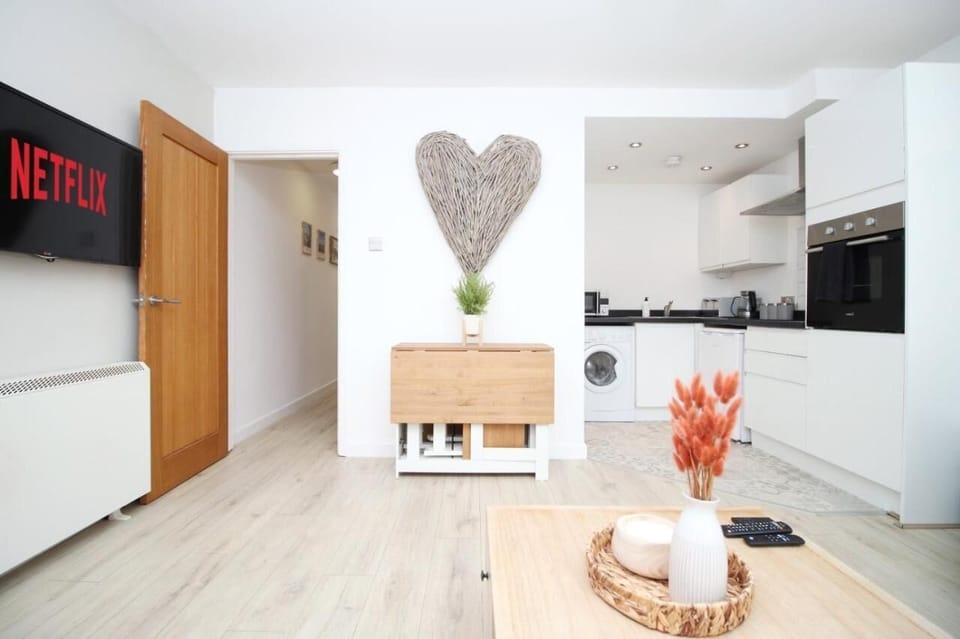 This inviting room blends comfort and practicality, featuring a mounted TV, decorative coffee table, and charming heart wall art. The open kitchen with sleek cabinetry and appliances ensures a seamless stay for guests.