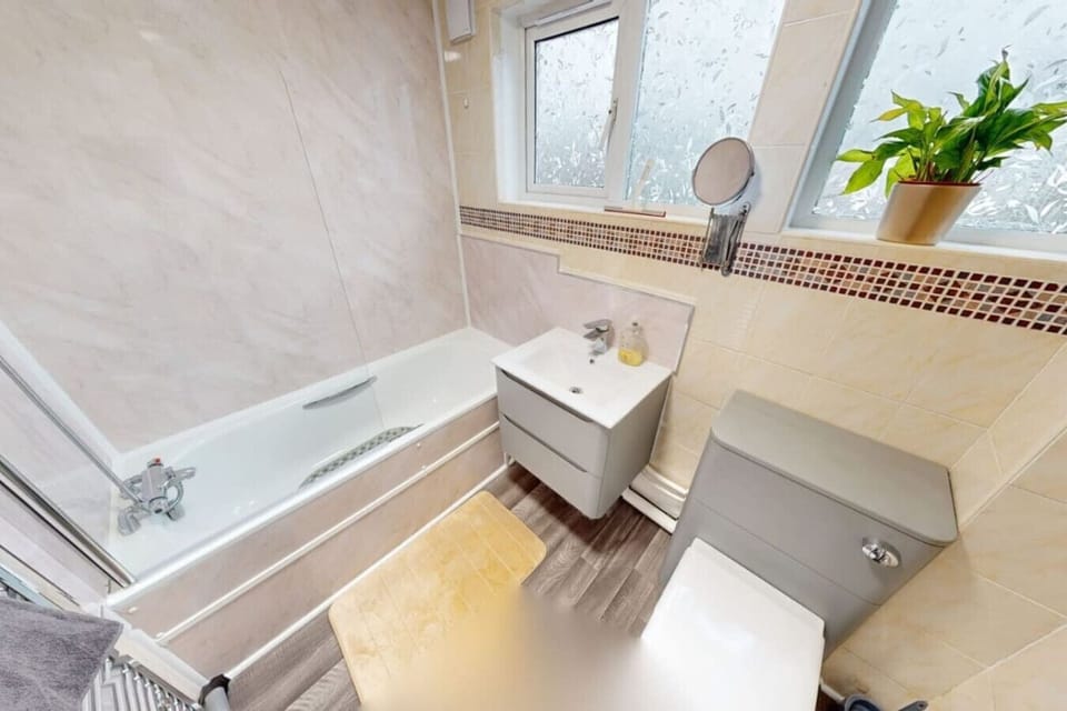 Tiled bathroom with a bath, overhead shower and glass screen, wall-hung vanity basin, toilet, mosaic tile border detail, and two frosted windows.