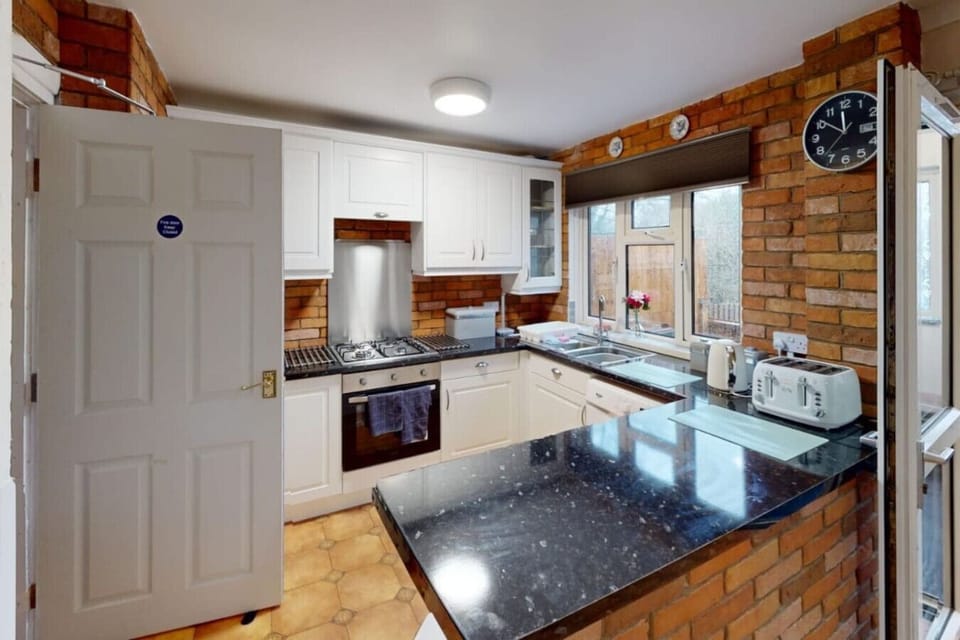 White fitted kitchen with a gas hob, built-in oven, dark worktop, exposed brick splashback, and countertop appliances including a kettle and toaster visible along the worksurface.