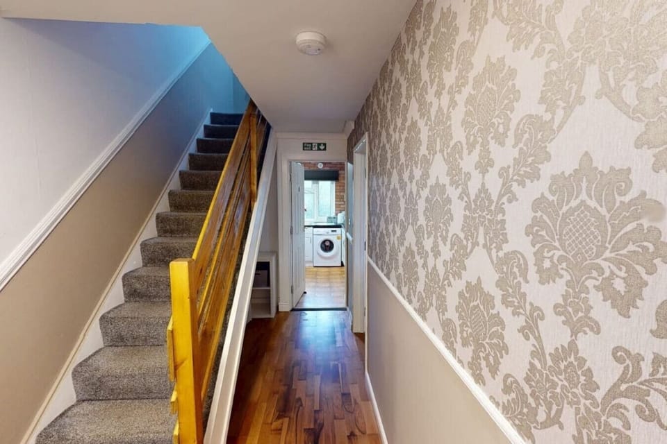 Carpeted staircase with a yellow-painted banister leading to an upper-floor landing, with a ground-floor corridor and a utility area containing a washing machine visible at the far end.