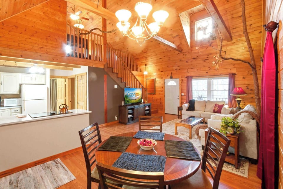A wide-angle view of the cabin's great room showcasing the round wooden dining table, a cozy living area with a plush sofa, and a staircase leading to the upper loft.
