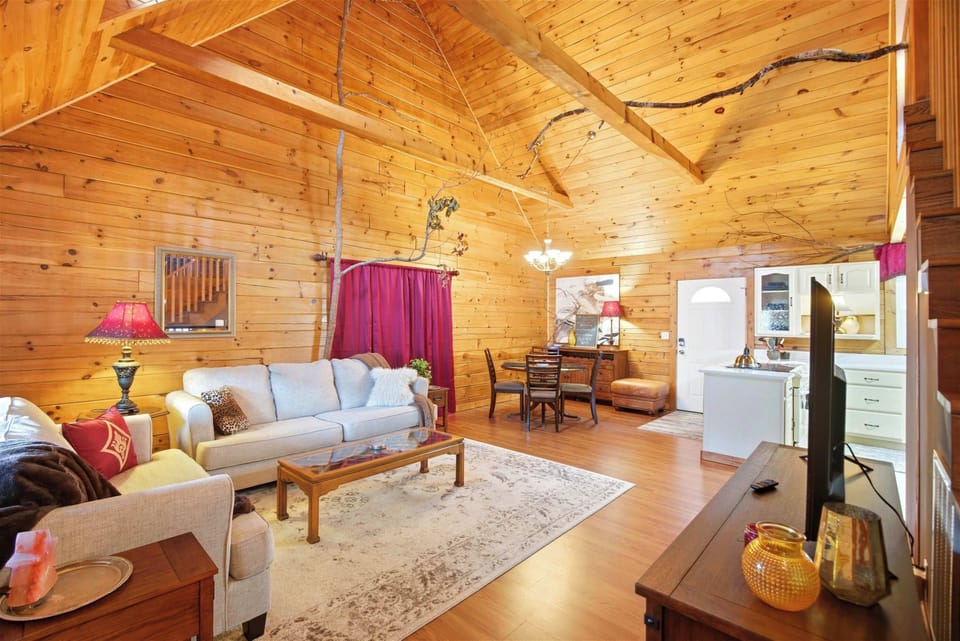 An open-concept living room in a log cabin featuring high vaulted ceilings, a large flat-screen TV on a wooden console, and a staircase leading to a loft sleeping area.