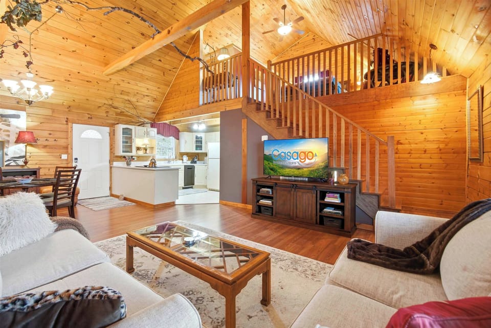 An open-concept living room in a log cabin featuring high vaulted ceilings, a large flat-screen TV on a wooden console, and a staircase leading to a loft sleeping area.