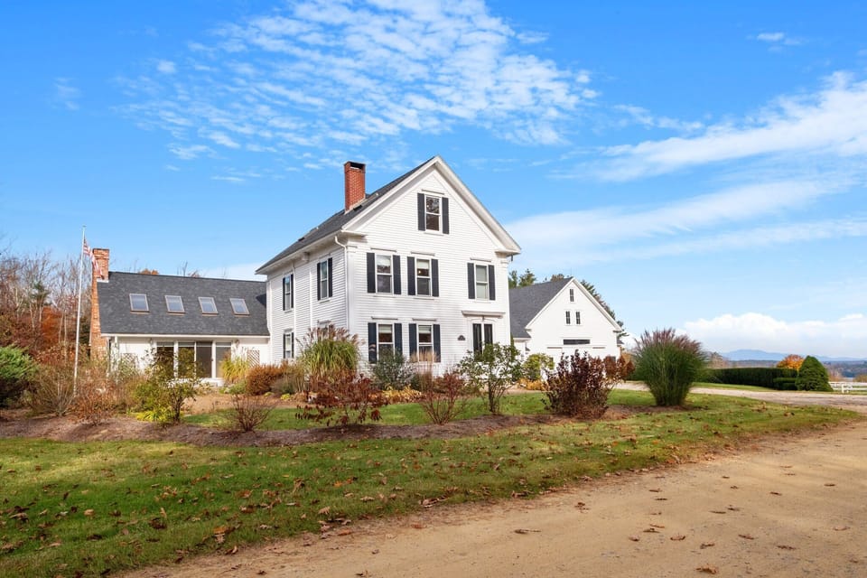 A classic white New England farmhouse set against open fields — your home base for exploring the Lakes Region.