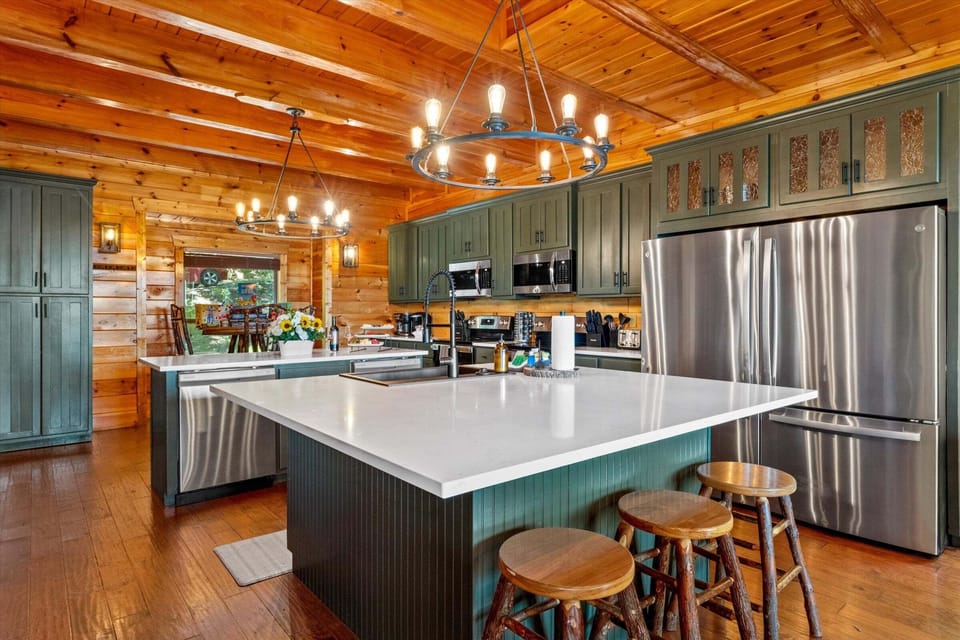 Large kitchen island with bar seating, stainless steel appliances, green cabinetry, wood ceiling, and an open dining area with mountain views.
