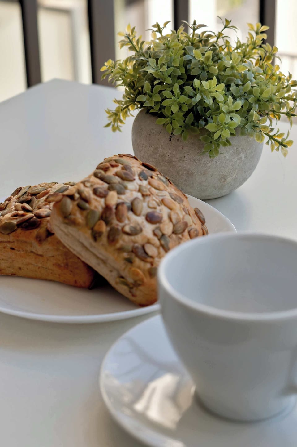 A table set with a plate of sliced bread and a mug is seen in this picture. Next to it is a vase with a small flowering plant, and in the background is a blurred window.