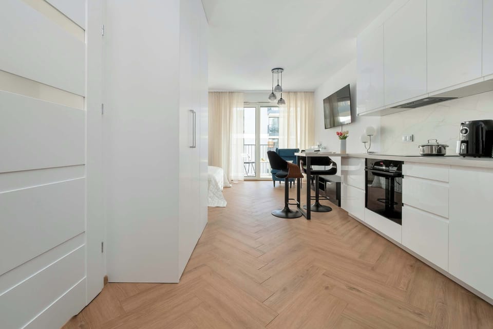 A spacious apartment hallway with wooden flooring and bright lighting. Large white kitchen counters can be seen on the right side of the photo.