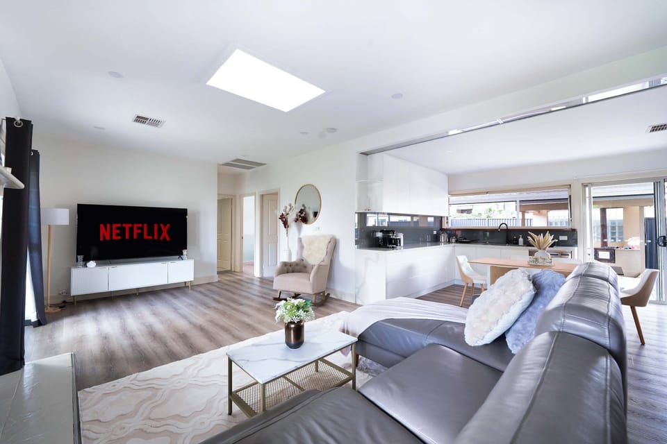 Bright, spacious living room with high ceilings, abundant natural light, garden views & classic fireplace. Features a 70” Smart TV with Netflix, electric reclining leather sofa, rocking chair, coffee table, stylish art, AC, and soft LED lighting.
