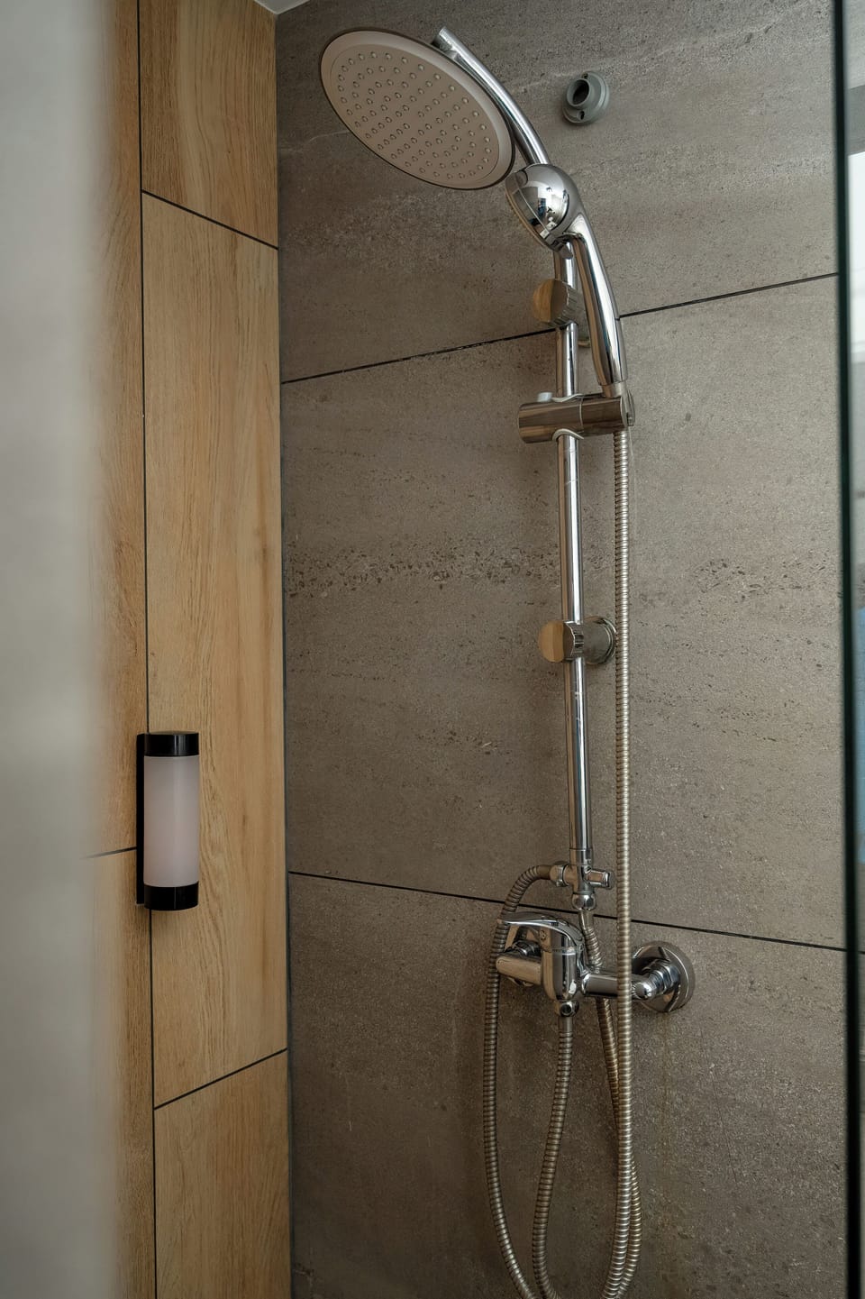 A shower area with a glass partition and a wall-mounted shower fixture. The walls are covered with modern gray tiles. The space looks clean and minimalistic.
