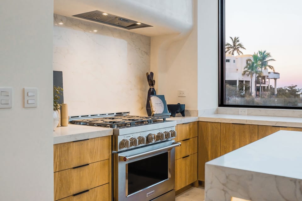 Your sleek kitchen features professional-grade appliances and warm wood cabinetry, with tropical palm views through the window creating a perfect cooking backdrop.
