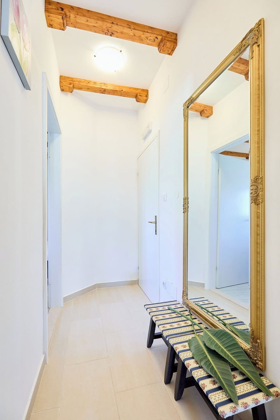 Step into a hallway where rustic charm meets elegant design. Exposed wooden beams grace the ceiling, complementing the clean, modern aesthetic and a stunning full-length gold mirror.